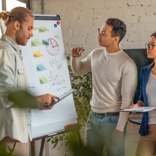 Young man standing by flipchart and giving presentation over new business startup to his collegues in modern office