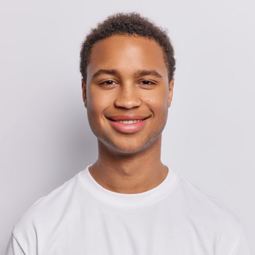 Portrait of pleased dark skinned man with short curly hair stands satisfied glad to hear good news dressed in casual t shirt isolated over white background. People and positive emotions concept